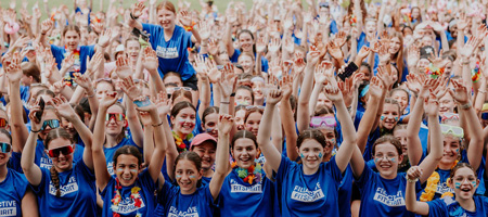 About 100 young girls in their teens have their arms raised straight up in a cheerful manner. 