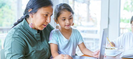 Two Indigenous females, a woman in her 50s and a 5-year-old girl are sitting in front of a laptop.  