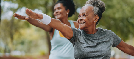 Two Afro-American women in their 70s are smiling while doing yoga in a park surrounded by trees. 